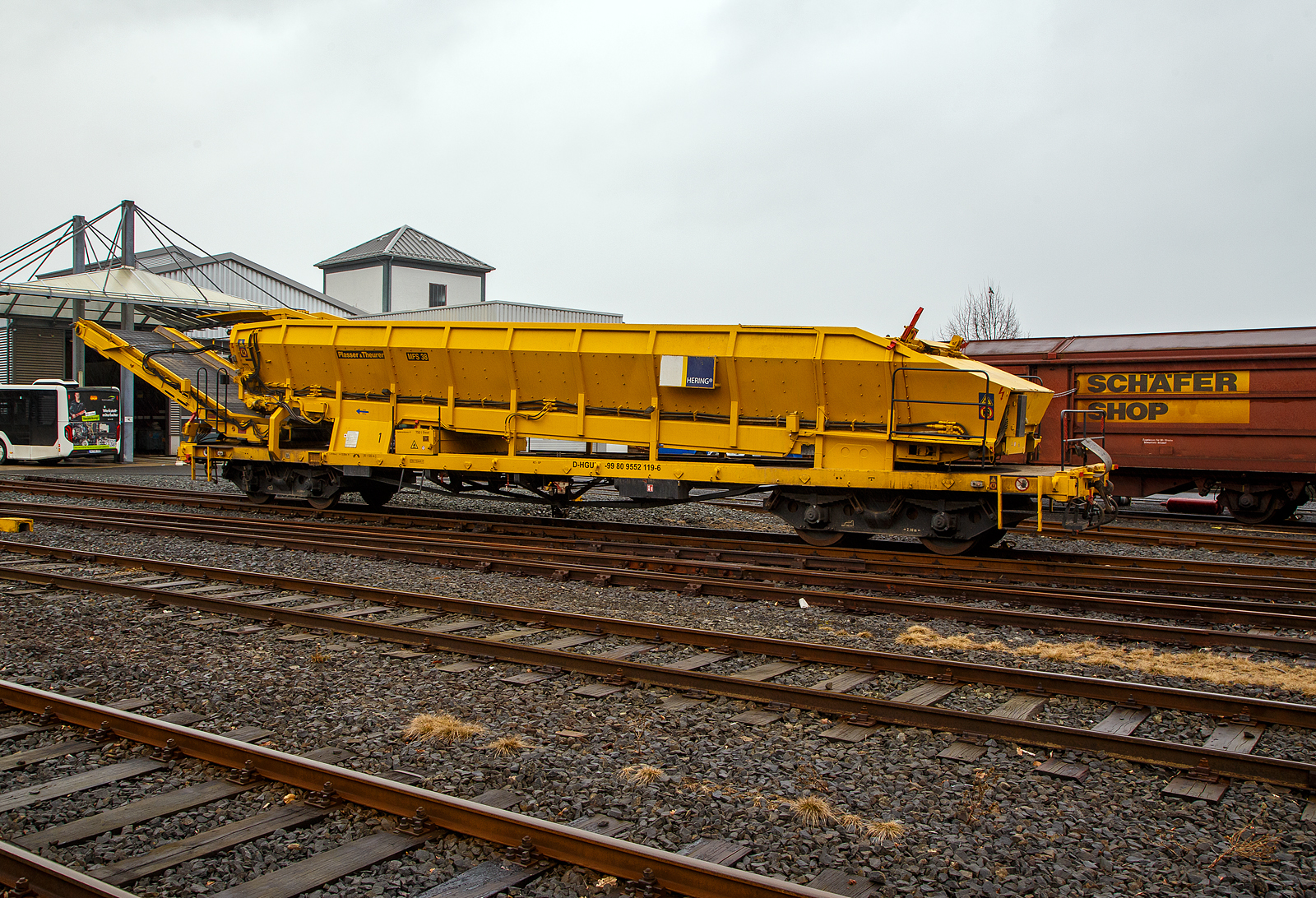 Plasser & Theurer Materialförder- und Siloeinheiten MFS 38 (teilweise auch BSW 2000 - Bunker-Schüttgut-Wagen genannt) der HERING Bahnbau GmbH, Burbach, Nr. 1, Schweres Nebenfahrzeug Nr. D-HGUI 99 80 9552 119-6, ex 97 19 22 552 17-1, ist am 13.03.2023 auf dem Betriebshof der Westerwaldbahn (WEBA) auf der Bindweide bei Steinebach/Sieg abgestellt.

Diese MFS 38 wurde 1993 von Plasser & Theurer unter der Fabriknummer 2159 gebaut, wobei als Fahrgestell ein vierachsiger ehemaliger Drehgestell-Flachwagen der Gattung Rs 680 (ex SSlmas 53), der 1962 von M.A.N. im Werk Nürnberg unter der Fabriknummer 146801 gebaut wurde, Verwendung fand. Diese Wagen wurden Plasser & Theurer vom Kunden beigestellt. 

Bei diesem MFS 38 (ohne nachfolgendes „D“) ist das Übergabeband beidseitig zum Gleis um 45 Grad schwenkbar, so ist ein  Entladen nebens Gleis oder zum Nachbargleis möglich. Die MSF 38 haben ein Fassungsvermögen 38 m³ bzw. eine Nutzlast von 42t, bei einem Eigengewicht von 38t. Die Förderbänder werden hydraulisch angetrieben. Der Antrieb des Hydraulikaggregates erfolgt durch einen luftgekühlten Deutz Dieselmotor vom Typ F6L 912 mit einer Leistung von 82 kW.

TECHNISCHE DATEN:
Maschinenart: Material-Förder-Siloeinheit MSF 38
Hersteller: Plasser & Theurer
Spurweite: 1.435 mm (Normalspur)
Anzahl der Achsen: 4 (in 2 Drehgestellen)
Länge über Puffer:19.900 mm
Gesamtlänge mit Übergabeband: 23.550 mm
Drehzapfenabstand: 13.000 mm
Achsabstand im Drehgestell: 2.000 mm
Laufraddurchmesser: 1.000 mm (neu)
Drehgestell-Bauart: Minden Dorstfeld-Drehgestell 931 / 932
Breite: 2.890 mm
Höhe: 4.280 mm
Eigengewicht: 38 t
Max. Zuladung (Nutzlast): 42 t
Ladevolumen: 38 m³
Höchstgeschwindigkeit: 100 km/h (geschleppt)
Kleister befahrbarer Gleisbogen: R=120 m (angeschrieben)
Bremse: KE – GP
Motor: luftgekühlter Deutz Dieselmotor vom Typ F6L 912 F
Leistung: 82 kW
Dieseltankvolumen: 750 l
Übergabeband: +/- 45 Grad schwenkbar.
Entladezeit: 3,5 Minuten
Entladeabstand von Gleismitte: max. 4,5 m
Die Förderbänder sind in folgenden Geschwindigkeiten regelbar:
Siloband: 1. Gang (Speichern)   0 - 0,028 m/s / 2. Gang (Durchfördern)  0 - 0,140 m/s
Übergabeband: 0 - 1,000 m/s


Einsatz und Arbeitsweise:
Bei diesem MFS 38 (ohne nachfolgendes „D“) ist das Übergabeband beidseitig zum Gleis um 45 Grad schwenkbar, so ist ein  Entladen nebens Gleis oder zum Nachbargleis möglich. Die MSF 38 haben ein Fassungsvermögen 38 m³ bzw. eine Nutzlast von 42t, bei einem Eigengewicht von 38t. Die Förderbänder werden hydraulisch angetrieben. Der Antrieb des Hydraulikaggregates erfolgt durch einen luftgekühlten Deutz Dieselmotor vom Typ F6L 912 mit einer Leistung von 82 kW.

Mit den Materialförder- und Siloeinheiten können unterschiedliche Schüttgüter wie beispielsweise Abraum, Planum- und Frostschutzmittel oder Bettungsstoffe unkompliziert, geladen, gefördert, gespeichert und entladen werden. Das Abraumverladesystem ist weitestgehend automatisiert und benötigt zur Bedienung nur wenig Personal. Das System verfügt zudem über eine autarke Energieversorgung (durch Dieselmotor). Ein unabhängiges Entladen ist durch schwenkbare Übergabebänder an jeder geeigneten Stelle möglich. Die MFS-Wagen können in Kombination mit einer Beladestation in beliebiger Anzahl aufgereiht und vor Kopf be- und entladen werden.

Am Boden jedes MFS 38 befindet sich ein über die ganze Wagenbreite gehender Fördergurt und an der Stirnseite ein breites Abgabeförderband. Das Aushubmaterial fällt von der Maschine kommend auf den Bodengurt, wird zum Abgabeförderband gebracht und an die nächste Einheit übergeben. Dieser Vorgang wiederholt sich, bis die am weitesten entfernte Siloeinheit erreicht wird. Bei dieser hintersten Siloeinheit ist das Abgabeförderband an der Stirnseite des Wagens in Ruhestellung. Auch der Fördergurt am Boden des Wagens ist so lange in Ruhestellung, bis die obere Füllgrenze erreicht ist. Dann wird der Fördergurt in Bewegung versetzt und dadurch im Übergabebereich freier Raum für die weitere Beladung geschaffen. Durch die dem Fülltempo angepasste Geschwindigkeit des Fördergurts wird die ganze Siloeinheit vollständig gefüllt. Das Fassungsvermögen einer Siloeinheit beträgt 38 Kubikmeter.

Nach Füllung einer Teilgruppe des Abraumzuges kann diese von der Restgruppe abgekuppelt werden, zu einer Entladestelle gefahren und dort entleert werden, während die Restgruppe des Abraumzuges noch befüllt wird. Das Entladeband ist nach beiden Seiten ausschwenkbar. Die Entladung kann dabei auf einer Deponie erfolgen oder es können LKW's oder Waggons am Nachbargleis beladen werden. Die MFS 38 (ohne nachfolgendes „D“) des Abraumzuges können durch Ausschwenken aller Abgabeförderbänder gleichzeitig entleert werden. Die Entladung eines Wagens dauert ungefähr 3,5 Minuten.