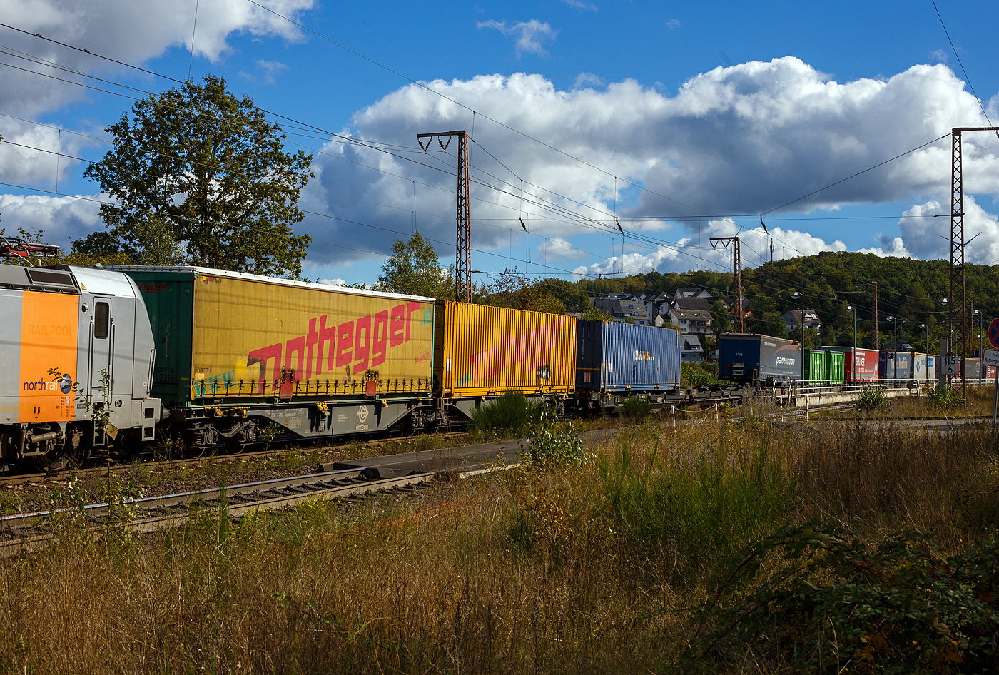 Sechsachsige Drehgestell-Gelenk-Containertragwagen-Einheit / 6-achsiger Containertragwagen, 33 87 4901 507-6 F-ERSA, der Gattung Sggmrss 90', der franz�sischen Vermietungsfirma ERMEWA S.A., am 05 Oktober 2024 im Zugverband, direkt hinter der Vectron AC193 922-2 der Northrail GmbH, im Zugverband bei der Zugdurchfahrt in Rudersdorf (Kr. Siegen). Hier beladen mit einer Wechselbr�cke und einem 45-Fu�-ISO-Containern.

Der Wagen der Gattung Sggmrss wird f�r den Transport von Containern und Wechselbeh�ltern verwendet und hat drei zweiachsige Drehgestelle. Die Flachwagen sind f�r 20-, 30-, 40- und 45-Fu�-ISO-Container und Wechselbeh�lter geeignet.

TECHNISCHE DATEN:
Spurweite: 1.435 mm (Normalspur)
L�nge �ber Puffer: 29.590 mm
Drehzapfenabst�nde: 2 x 11.995 mm
Achsabstand in den Drehgestellen: 1.800 mm
Laufraddurchmesser: 920 mm (neu)
Ladel�nge: 2 x 13.820 mm
H�he der Ladeebene �ber S.O.: 1.155 mm
Eigengewicht: 27.480 kg
Max. Zuladung bei Lastgrenze S: 107,5 t (ab Streckenklasse D)
Max. Zuladung bei Lastgrenze SS: 92,5 t (ab Streckenklasse C)
H�chstgeschwindigkeit: 100 km/h Lastgrenze S / 120 km/h bei Lastgrenze SS und leer)
Kleinster befahrbarer Gleisbogen: R 75 m
Bremse: 2 x KE-GP-A (K)
Bremssohle: Cosid 810
Verwendungsf�higkeit: TEN GE / G1
