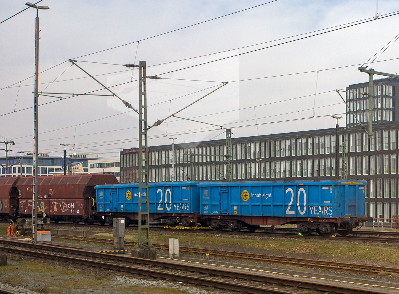 Sieht aus als seien es zwei vierachsiger offener Drehgestell-Güterwagen (Hochbordwagen) der Gattung Eanos, ist es aber nicht, denn es ist:
Ein 2 x 4-achsiger Flachwagen in Sonderbauart (Doppelwagen) bzw. ein 8-achsiger Containertragwagen, 31 81 485 4 229-9 A-IF, der Gattung Sggrrs
80 ft, der österreichischen INNOFREIGHT hier als Wagon der Variante Q – mit zwei aufgesetzten ScrapTainer mit einer Länge von je 40' der DB Cargo AG, abgestellt am 10 März 2024 beim Hauptbahnhof Oldenburg (Oldb). Aufnahme aus einem Zug herraus.

Grundsätzlich besteht ein InnoWaggon aus zwei kurzgekuppelten Einzelwagen. Leichtbauweise und hohe Belastbarkeit differenzieren diese Waggons dabei klar von anderen und ermöglichen deutlich höhere Zuladung für den Transport von unterschiedlichsten Gütern. Der InnoWaggon vereint den Tragwagen und die auswechselbaren Behälter zu einem innovativen Konzept, das eine flexible Nutzung verspricht. Der Einsatz der Spezialbehälter wird dabei der jeweiligen Branche angepasst, da die Behälter je nach Anforderung wie spezifisches Schüttgewicht oder Entladestation eingesetzt werden.

Der Wagen wurde 2016 Tatravagonka Poprad (Slowakei) gebaut.

TECHNISCHE DATEN des 2x40 ft InnoWaggon:
Gattung: Sggrrs
Gattungskennzah:l 4854
Spurweite: 1.435 mm (Normalspur)
Achsenanzahl : 8 pro Doppelwagen (in 4 Drehgestellen)
Länge über Puffer : 26.710 mm (mit Langhub-Puffer)
Breite: 2.878 mm
Drehzapfenabstand: 2 x 8.070 mm
Achsabstand in den Drehgestellen: 1.800 mm
Raddurchmesser: 920 mm (neu)
Länge Ladefläche: 2 x 12.370 mm (2 x 40 ft)
Höhe der Ladefläche über SOK: 1.155 mm
Eigengewicht : ca. 29,700 kg
Max. Geschwindigkeit: 100 km/h (beladen) / 120 km/h (leer)
Max. Zuladung pro Doppelwagen: 150,3 t (ab Streckenklasse D, ohne Behälter)
Genaue Lastgrenzen nach Ausführungsvariante (Lastgrenzenraster Stecktafel)
Max. Achslast 22,5 t
Ladequerschnitt: 8 m²
Ladelänge: 28.130 mm (90 ft)
Ladegut: Rundholz in 4 oder 5m Länge
Lademöglichkeit: 6 x 4 m Pakete oder 5 x 5 m Pakete
Entlademöglichkeiten: Logstacker oder Highlifter
Bremse: KE-GP-A (K)
Bremssohle: Jurid 822
Kleinster befahrbarer Gleisbogen: R = 75m
Intern. Verwendungsfähigkeit: TEN-GE

Die Container (ScrapTainer):
Bei dem neu von Innofreight entwickelten ScrapTainer handelt es sich um einen offenen Container für den Transport von Eisen-, Stahl- und Mischschrott, schwere Schüttgüter, sowie für Kohle- und Kokstransporte. Die ersten wurden im Oktober 2019 geliefert. Zusammen mit dem ebenfalls von Innofreight entwickelten 80-Fuß InnoWaggon ergeben die Behältnisse ein Eigengewicht von 43,5 Tonnen und eine maximale Zuladung von 136,5 Tonnen Fracht.

Im regulären Einsatz ist zum Zwecke der Entladung kein Abnehmen des Behälters vom Waggon vorgesehen, das Ladegut wird mittels Kran oder Bagger entleert. Eine Abnahme der Container ist jedoch für Instandhaltungs- oder Lagerzwecke möglich, weshalb der Wagen auch universell für sämtliche weitere Aufbauten verwendet werden kann.

TECHNISCH DATEN eines ScrapTainer:
Länge: 40 f
Ladelänge: 12.100 mm
Ladevolumen: 70,1 m³
Ladefläche: 33,2 m²

DB Cargo mietet von der deutschen Innofreight-Niederlassung einen ScrapTainer-Zug für Schüttguttransporte zwischen Deutschland und Österreich. Innofreight Solutions mit Sitz in Bruck an der Mur ist spezialisiert auf Entwicklung und Bau von innovativen, patentierten Gütertransport- und Logistiksystemen. Europaweit sind 12.000 Container und 1.200 InnoWaggons bei namhaften Bahngesellschaften im Einsatz.

Quellen: INNOFREIGHT und Anschriften