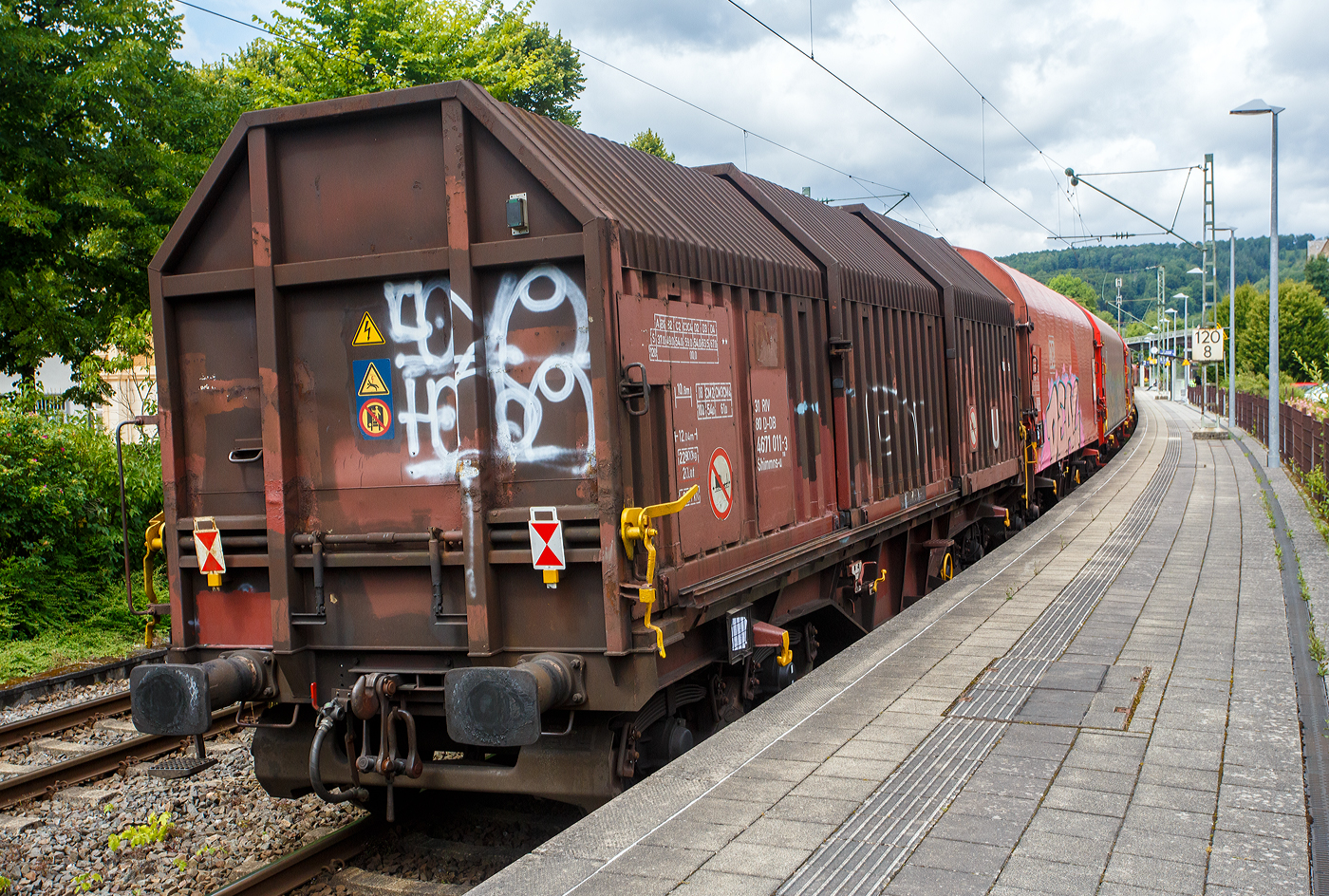 Vierachsiger Drehgestell-Flachwagen mit verschiebbaren Teleskophauben für Coiltransporte, 31 80 4671 011-3 D-DB, der Gattung Shimmns-u 708, der DB Cargo AG, am 16 Juli 2025 im Zugverband (Zug 9921) bei der Zugdurchfahrt in Kirchen (Sieg) in Richtung Siegen bzw. Ziel Kreuztal. 

Der Wagen eignet sich speziell für den Transport witterungsempfindlicher Blechrollen. Er besitzt feste Stirnwände, 5 fest im Untergestell eingebaute Lademulden und drei teleskopartig angeordnete Hauben, die sich zu beiden Wagenenden hin soweit durch Ineinanderschieben öffnen lassen, dass jeweils 2/3 des Wagens freigelegt werden kann. Hierdurch ist eine einfache Beladung des Wagens sowohl von oben als auch von der Seite aus möglich. Der Wagen darf nur mit geschlossenen und verriegelten Hauben verkehren.

Das Untergestell und die festen Stirnwände, die als Abkantrahmenkonstruktion mit Blechbekleidung und hutförmigen Verstärkungsprofilen ausgeführt sind, sowie die fest auf den Außenlangträgern angeordneten drei Laufschienen je Wagenseite bilden den als Schweißkonstruktion ausgeführten Wagenkasten. Das Untergestell mit seinen beiden kräftigen Außenlangträgern besteht aus Walz- und Abkantprofilen. Die Untergestellenden bestehen aus den Hauptquerträgern, den AK-Trägern und den Kopfstücken. Die sattelförmig angeordneten, jeweils mit den Außenlangträgern verschweißten Querträger bilden zusammen mit 6 mm dicken Muldenblechen die Lademulden.

Die teleskopartig ineinander verschiebbaren Stahlhauben bestehen aus einer Hohlprofil-Rahmenkonstruktion und einer mit Sicken versehenen Beblechung. Die Abdichtung der Hauben untereinander und zu den Stirnwänden hin erfolgt über labyrinthartig ausgeführten Umrandungsprofilen. Die Endhauben verriegeln in geschlossener Stellung automatisch. Das Entriegeln der Hauben ist sowohl vom Erdboden wie auch von der Rampe möglich. Die beidseitig an jeder Stirnwand angeordneten Verriegelungseinrichtungen sind durch Wellen miteinander verbunden. Hierdurch ist das Entriegeln von nur einer Wagenseite aus möglich. Jede Haube läuft auf 4 jeweils und mit Abhebesicherungen versehenen Laufwagen mit nadelgelagerten Doppelrollen.

Die Wagen der Bauart Shimmns 708 sind mit 12 verstellbaren Festlegearmen ausgerüstet, die in fest auf den Muldenkappen angeordneten kammartig ausgeführten Halterungen gelagert sind. Die Festlegearme und ihre Anordnung ergeben einen wirksamen Schutz gegen unzulässiges seitliches Verschieben der Coils.

Um unzulässige Radlastabweichungen auszuschließen, ist eine möglichst mittige Verladung der Coils erforderlich. Ein durch alle Mulden in Wagenmitte verlaufender weißer Farbstreifen dient hierzu als Orientierungshilfe.

TECHNISCHE DATEN:
Hersteller: LHB - Linke-Hofmann-Busch GmbH in Salzgitter-Watenstedt
Baujahr: 1972, Fabriknummer 407
Spurweite: 1.435 mm (Normalspur)
Achsanzahl: 4 (in 2 Drehgestelle)
Länge über Puffer: 12.040 mm
Länge der Ladefläche: 10.800 mm
Ladebreite (Mulden): 2.400 mm
Drehzapfenabstand: 7.000 mm
Achsabstand im Drehgestell: 1.800 mm
Laufraddurchmesser: 920 mm (neu)
Eigengewicht: 22.800 kg
Höchstgeschwindigkeit: 100 km/h (beladen) / 120 km/h (leer)
Maximales Ladegewicht: 67,0 t (ab Streckenklasse D4) / 61,0 t bei DB ab CM3
Hier betrug das Ladegewicht laut Laufzettel 65.055 kg
Kleinster bef. Gleisbogenradius: 35 m
Bauart der Bremse: KE-GP (LL)
Bremssohle: IB 116
Handbremse: Ja
Intern. Verwendungsfähigkeit: RIV