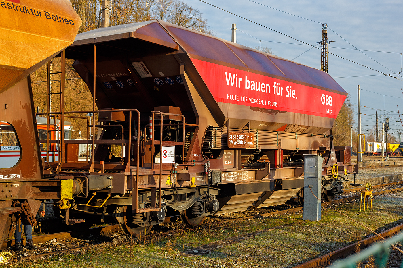 Vierachsiger offener selbstentladender Schotterwagen mit einfacher hydraulischer Bedienung, 31 81 6985 014-2 A-�BB, der Gattung Faccns, der �BB Infra, abgestellt am 14 Januar 2025 beim Bahnhof Lambach (Ober�sterreich).

Dieser 4-achsiger G�terwagen Faccns ist ein selbstentladender Schotterwagen, der mit 4 Drehschiebern ausgestattet ist. F�r den Entladevorgang wird die
Schwerkraft des Ladeguts genutzt. Vier Auslauf�ffnungen mit Drehschiebern
erm�glichen eine restlose Entladung des Wagens auf beiden Seiten, zwischen oder neben den Schienen. Der Wagen ist f�r den Transport nicht n�sseempfindlicher G�ter in loser Sch�ttung geeignet. Wagen ist mit hydraulischer Bet�tigung der Auslaufrutschen und den Schiebern ausger�stet. Der Vorteil besteht darin, dass dieser Wagen keine externe Energiequelle braucht. Druck wird durch das Fahren mit Hilfe der Radsatzpumpen aufgebaut.Die Bet�tigungspanele des Hydrauliksystems sind von der Betriebsb�hne des Wagens zug�nglich.

Bedeutung der Gattungs- und Kennbuchstaben:
F = Offener Wagen in Sonderbauart
a = Mit 4 Rads�tzen
cc = mit Schwerkraftentladung, dosierbar, wahlweise zweiseitig, tiefliegend
n = mit vier Rads�tzen h�chste Lastgrenze �ber 60 t
s = H�chstgeschwindigkeit 100 km/h (beladen nach Lastgrenzraster)

TECHNISCHE DATEN der Faccns:
Hersteller: �OS Trnava a.s. (Slowakei)
Spurweite: 1.435 mm (Normalspur)
Anzahl der Achsen: 4 (in 2 Drehgestellen)
L�nge �ber Puffer: 12.540 mm
Drehzapfenabstand: 7.500 mm
Laufraddurchmesser: 920 mm (neu)
Laderaum:	 45,0 m�
Eigengewicht: 24.000 kg 
H�chstgeschwindigkeit: 100 km/h (beladen gem. Lastgrenzraster) / 120 km/h (leer)
Max. Ladegewichte: 66,0 t ab Streckenklasse D3
Kleinster bef. Gleisbogenradius: R = 60 m
Bremse: MH-GP-A (K)
Intern. Verwendungsf�higkeit: RIV